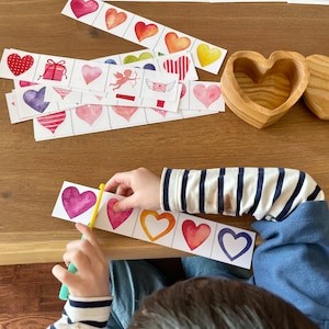 May include: A child is playing a matching game with colorful heart-shaped cards. The cards feature different designs, including hearts, gifts, and angels. The child is using a pair of scissors to cut out the hearts.