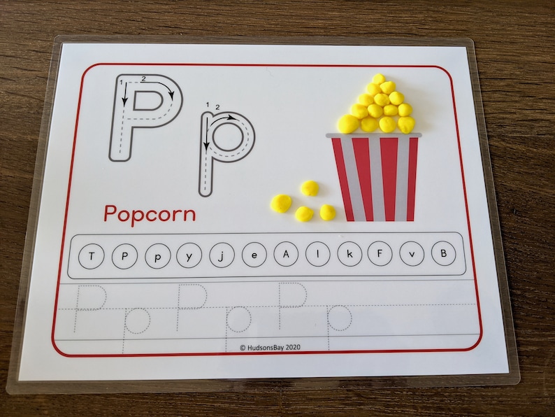 May include: A white learning card with a dotted line tracing the letter "P" and the word "Popcorn". The letter "P" is also formed with yellow pom-poms in a red and white striped container. There are also yellow pom-poms scattered below the container.