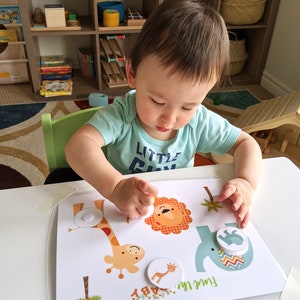 May include: A child sits at a table playing with a colorful animal matching game. The game features a giraffe, lion, and elephant with matching puzzle pieces. The game is labeled "Fun at the Zoo!"
