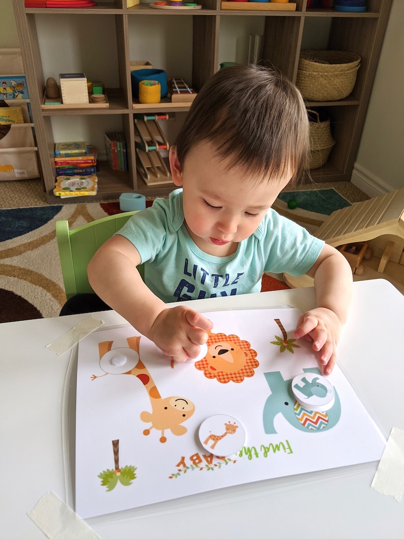 May include: A child sits at a table playing with a colorful animal matching game. The game features a giraffe, lion, and elephant with matching puzzle pieces. The game is labeled "Fun at the Zoo!"