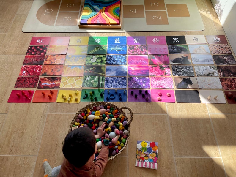 May include: A child plays with colorful wooden blocks and cards featuring images and Chinese characters for different colors. The cards are arranged in a rainbow pattern, with red, orange, yellow, green, blue, indigo, and violet. The child is sitting on the floor and reaching into a basket of colorful wooden blocks.