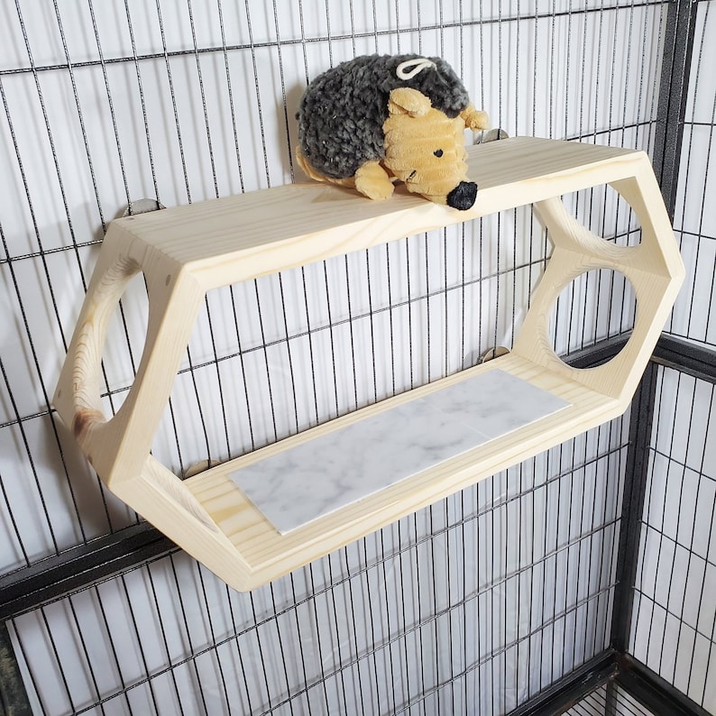 Puede incluir: Una estanter&iacute;a hexagonal de madera con una superficie de m&aacute;rmol. La estanter&iacute;a est&aacute; unida a una jaula de alambre negro y tiene un peque&ntilde;o juguete de peluche de erizo en la parte superior.