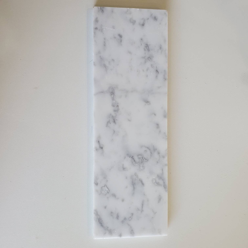 Puede incluir: Una baldosa de m&aacute;rmol blanco con vetas grises. La baldosa es rectangular y tiene una superficie lisa.