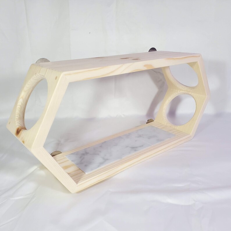 Puede incluir: Una estanter&iacute;a hexagonal de madera con una parte superior de m&aacute;rmol y dos recortes circulares. La estanter&iacute;a est&aacute; fijada a la pared con dos soportes plateados.