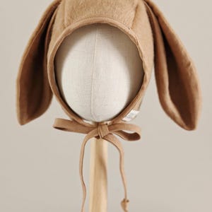 Könnte beinhalten: Ein beigefarbener Babyhut mit langen, schlaffen Hasenohren. Die Kapuze ist mit einer Schleife unter dem Kinn gebunden und wird auf einem Schaufensterpuppenkopf präsentiert. Der Hut ist aus einem weichen, flauschigen Material gefertigt.