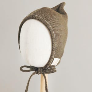 May include: A ribbed olive green bonnet featuring a pointed top, a white stitched trim along the face opening, and matching fabric ties that fasten under the chin. A small white rectangular brand label is attached to the side of the bonnet.