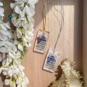 May include: Two rectangular resin pendants with a butterfly design and text, suspended on gold and silver chains. The pendants are displayed on a wooden surface, next to white flowers and a gold seashell.