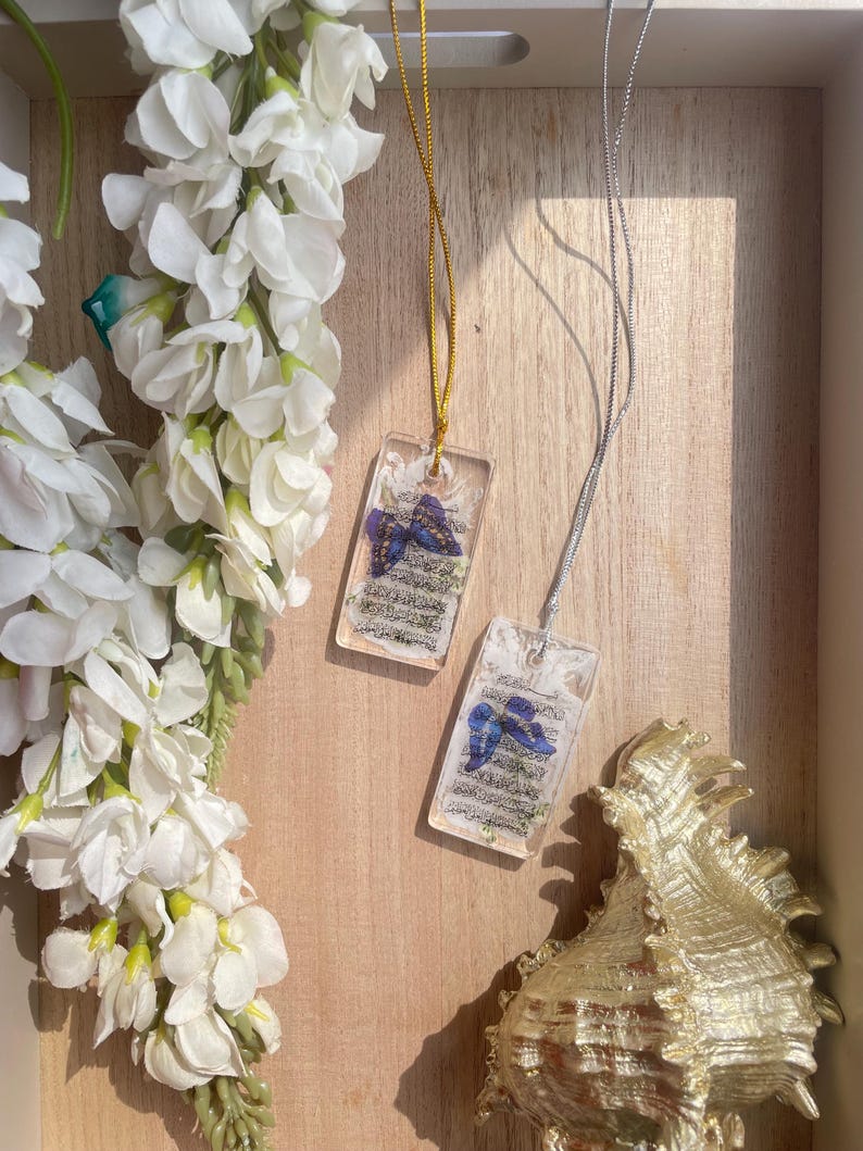 May include: Two rectangular resin pendants with a butterfly design and text, suspended on gold and silver chains. The pendants are displayed on a wooden surface, next to white flowers and a gold seashell.