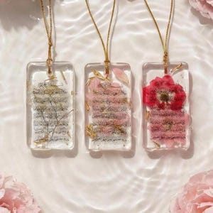 May include: Three rectangular resin pendants with gold chains. Each pendant features dried flowers and script. One pendant has a red flower, another has pink flowers, and the third has dried greenery.