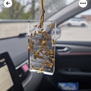 May include: A clear, rectangular resin car charm with gold leaf and Arabic script. It hangs from a gold cord inside a car. The background is blurred, showing a street scene. The charm is a decorative item.