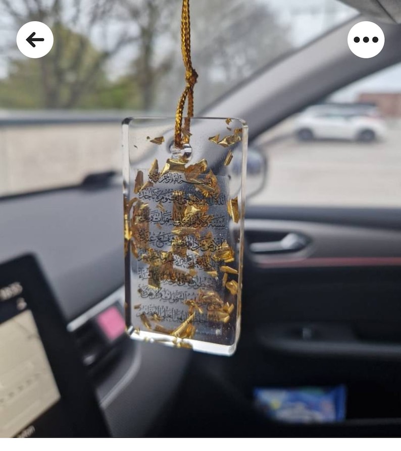 May include: A clear, rectangular resin car charm with gold leaf and Arabic script. It hangs from a gold cord inside a car. The background is blurred, showing a street scene. The charm is a decorative item.