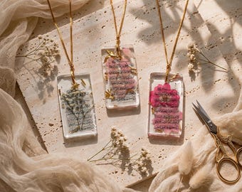 May include: Three rectangular resin pendants on gold-tone chains. Each pendant contains pressed flowers and sheet music fragments. The left pendant features white baby's breath, the centre has pink petals, and the right has a vibrant pink flower.