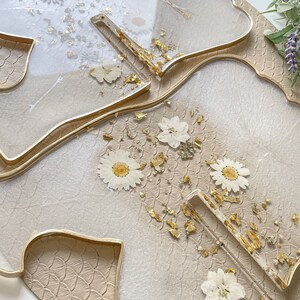 May include: Two clear resin trays with gold leaf and pressed white flowers. The trays are shaped like leaves and have a gold rim. The trays are on a beige textured surface.