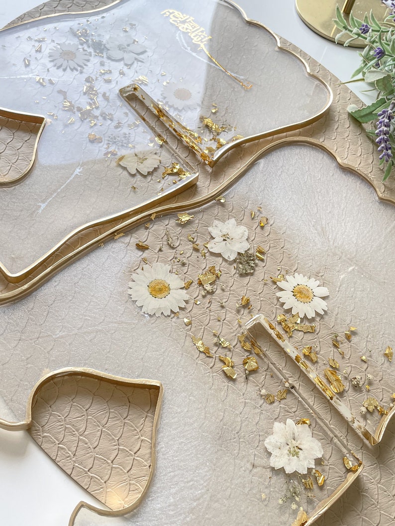 May include: Two clear resin trays with gold leaf and pressed white flowers. The trays are shaped like leaves and have a gold rim. The trays are on a beige textured surface.
