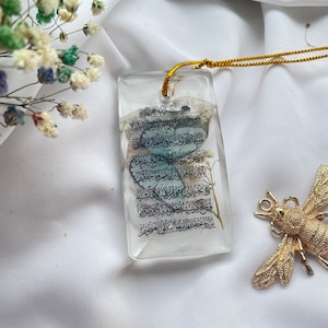 May include: A clear, rectangular pendant with a gold cord, featuring Arabic script and embedded dried flowers. A gold-coloured dragonfly-shaped brooch rests nearby. The pendant is set against a white fabric background.