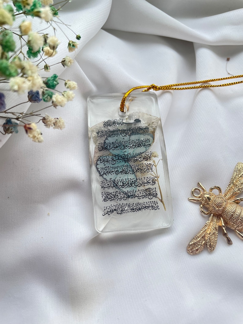 May include: A clear, rectangular pendant with a gold cord, featuring Arabic script and embedded dried flowers. A gold-coloured dragonfly-shaped brooch rests nearby. The pendant is set against a white fabric background.