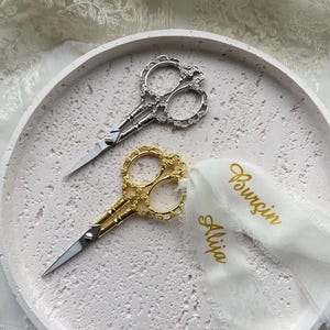 Könnte beinhalten: Zwei dekorative Scherenpaare, eine silberne und eine goldene, liegen auf einem hellrosa, runden Tablett. Die Scheren haben verzierte Griffe. Ein weißes Band mit den Namen "Alija" und "Burçin" in goldener Schrift ist an der goldenen Schere befestigt.