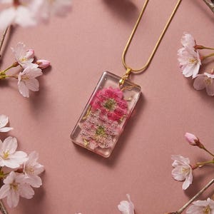 May include: A gold necklace featuring a rectangular pendant with pink flowers and text encased in clear resin. The background is a dusky pink, decorated with cherry blossom branches.