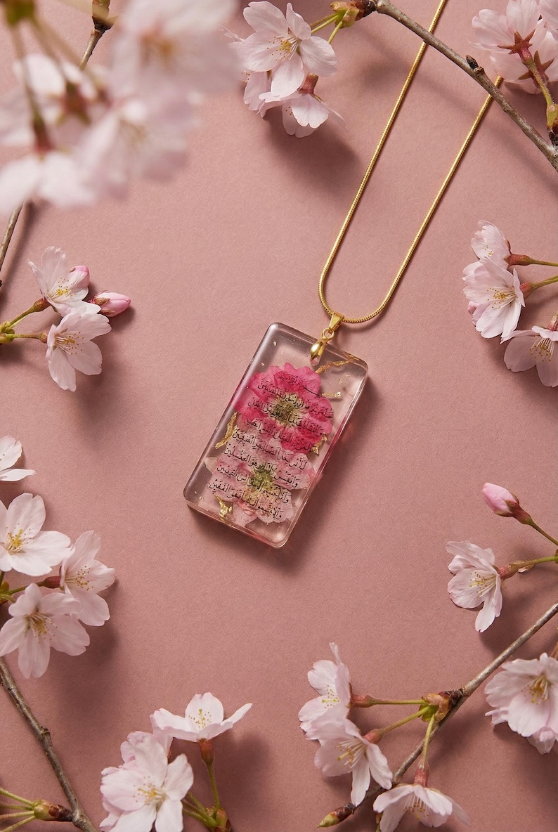 May include: A gold necklace featuring a rectangular pendant with pink flowers and text encased in clear resin. The background is a dusky pink, decorated with cherry blossom branches.