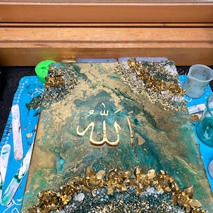 Könnte beinhalten: Ein abstraktes Gemälde in Grün und Gold mit goldenem Glitzer und Kristallen. Das arabische Wort "Allah" ist in Gold in der Mitte des Gemäldes geschrieben.