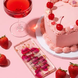 May include: A pink heart-shaped cake decorated with cherries and a glass of red liquid. A clear phone case with pink flower petals and gold flakes is next to strawberries on a pink surface.