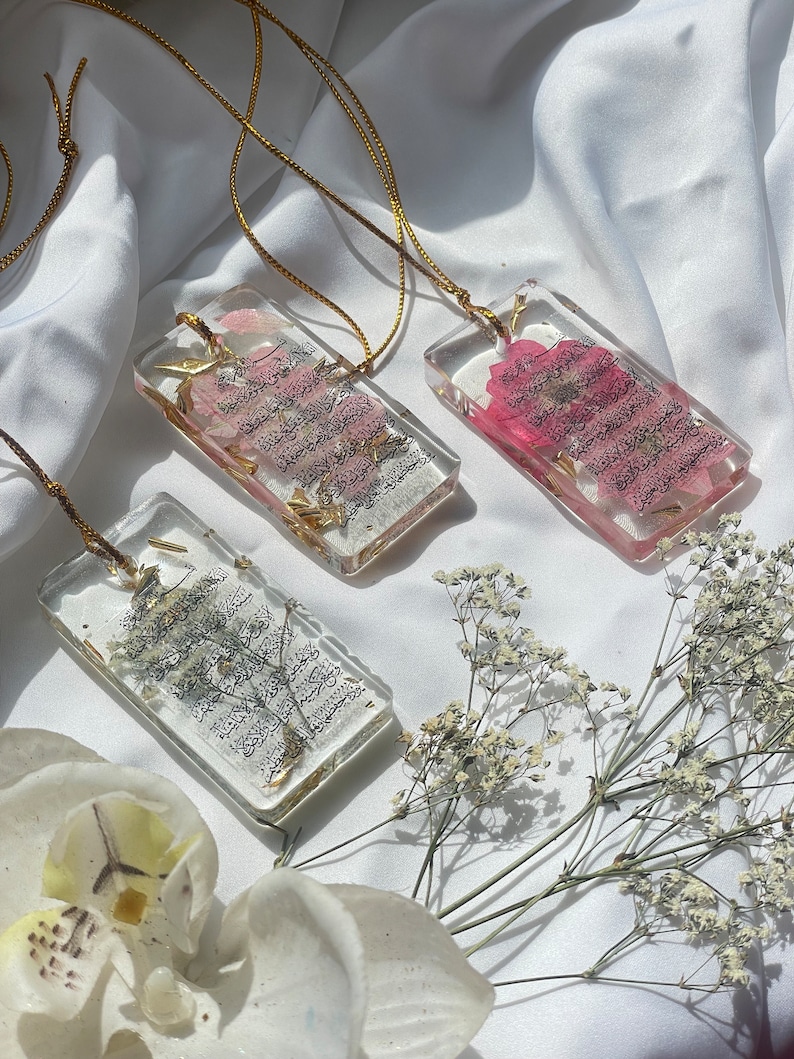 May include: Three rectangular resin pendants with gold-coloured cords. Each pendant contains dried flowers and script. The flowers are pink and white. The script is black. The pendants are on a white satin background.