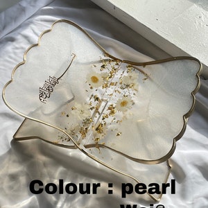 May include: A white resin book stand with a gold trim and a floral design. The stand is shaped like a wave and has the word "الله" written on it. The stand is labeled "Colour: pearl weiss".
