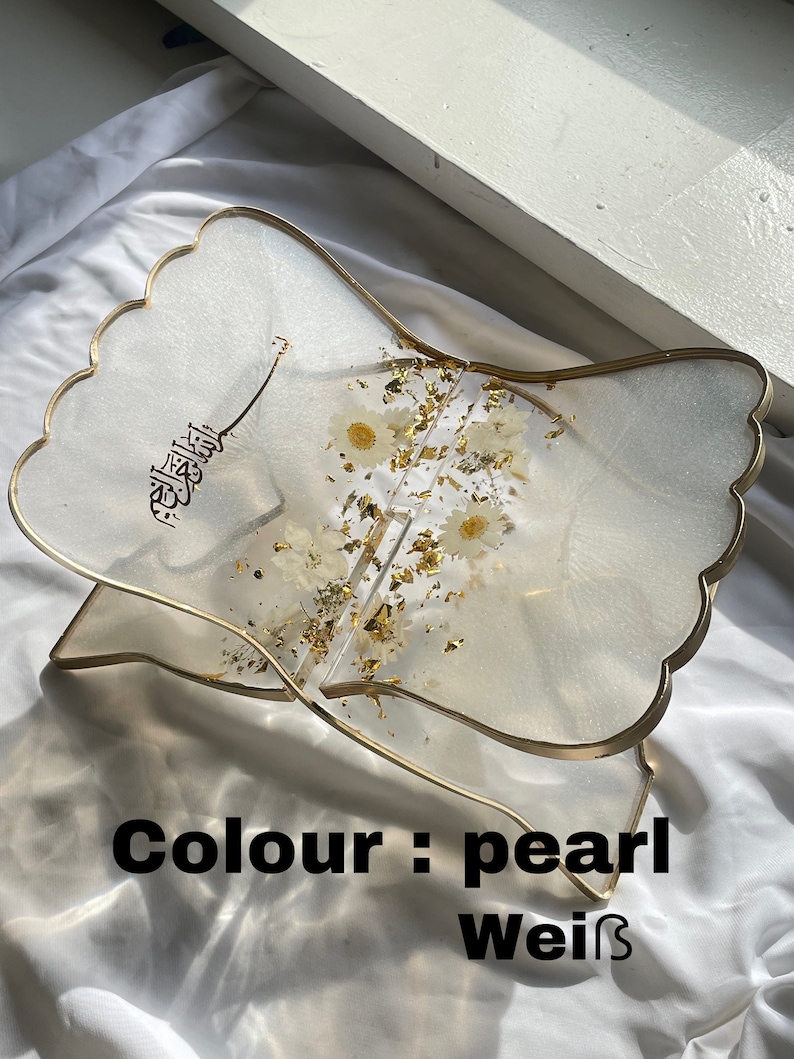 May include: A white resin book stand with a gold trim and a floral design. The stand is shaped like a wave and has the word "الله" written on it. The stand is labeled "Colour: pearl weiss".
