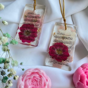 May include: Two rectangular resin pendants with gold cords, each containing a red flower and gold flakes. The pendants feature Arabic script. Pink and light pink flower-shaped soaps and a gold-coloured charm are also visible.