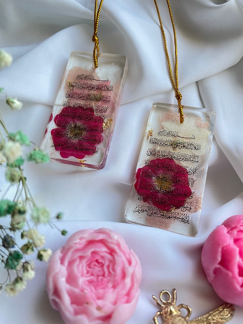 May include: Two rectangular resin pendants with gold cords, each containing a red flower and gold flakes. The pendants feature Arabic script. Pink and light pink flower-shaped soaps and a gold-coloured charm are also visible.