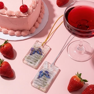 May include: Two rectangular pendants with butterfly designs and text, suspended on gold and silver cords, are displayed on a pink surface. A heart-shaped pink cake with cherries, strawberries, and a glass of red wine complete the scene.