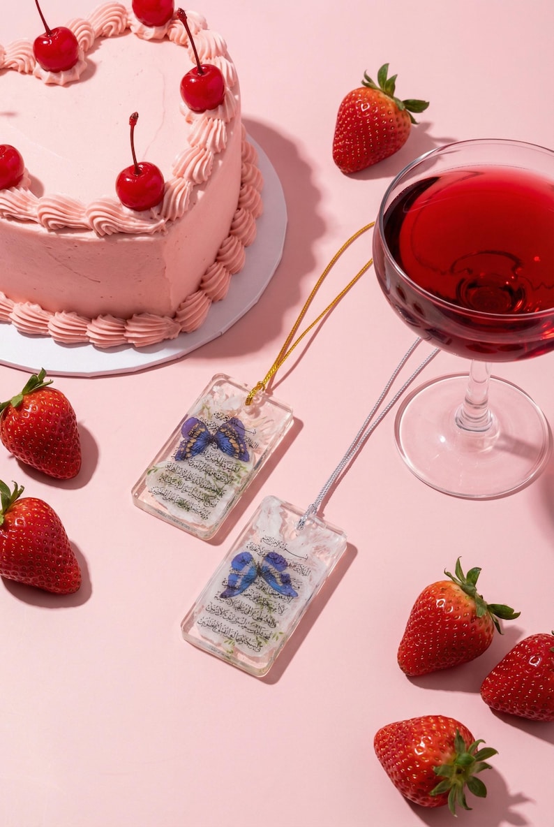 May include: Two rectangular pendants with butterfly designs and text, suspended on gold and silver cords, are displayed on a pink surface. A heart-shaped pink cake with cherries, strawberries, and a glass of red wine complete the scene.