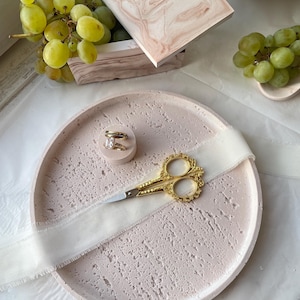 May include: A light pink textured tray with a white ribbon and gold scissors. Two gold rings sit on a small pink cylinder. A wooden box with a pink marble design and green grapes are also present.