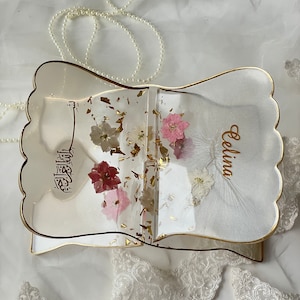 May include: A clear resin book with gold trim and a scalloped edge. The book is filled with dried flowers and gold flakes. The name "Celina" is written on the front cover.