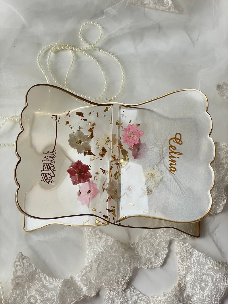 May include: A clear resin book with gold trim and a scalloped edge. The book is filled with dried flowers and gold flakes. The name "Celina" is written on the front cover.