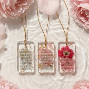 May include: Three rectangular resin pendants with gold chains, each containing dried flowers and script. The pendants are in clear resin, with a gold chain. The background features pink flowers and water.