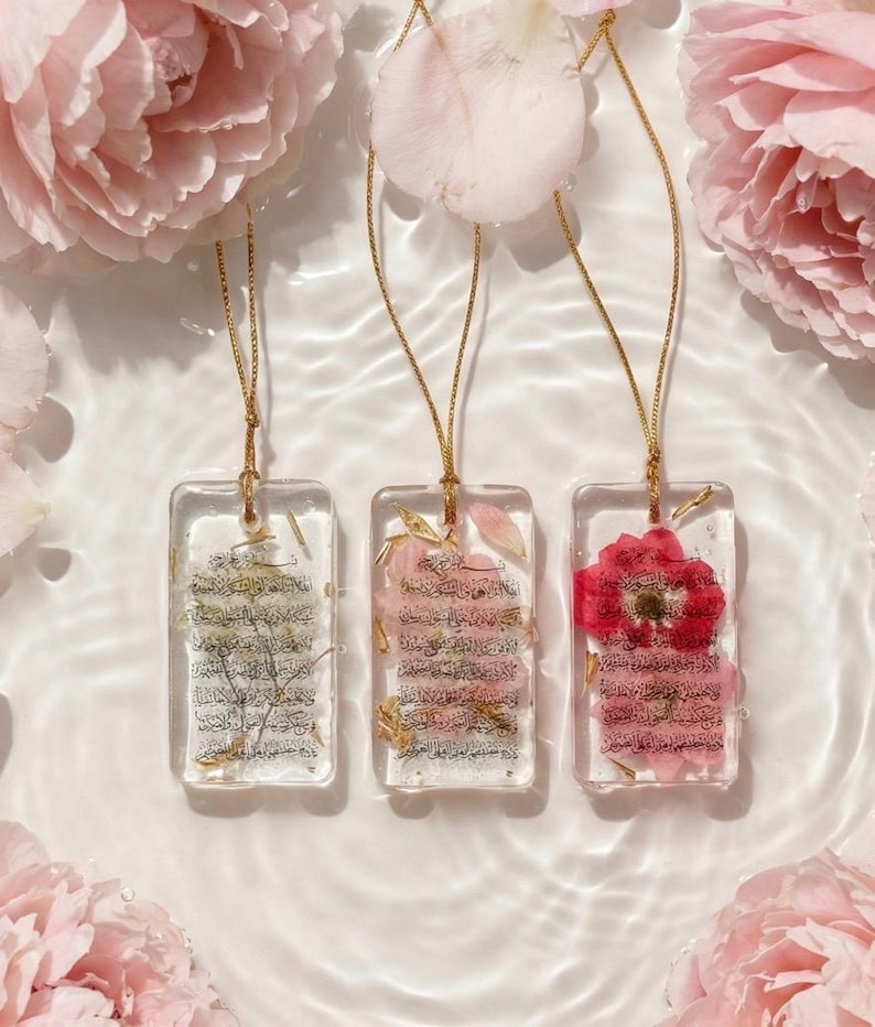 May include: Three rectangular resin pendants with gold chains, each containing dried flowers and script. The pendants are in clear resin, with a gold chain. The background features pink flowers and water.