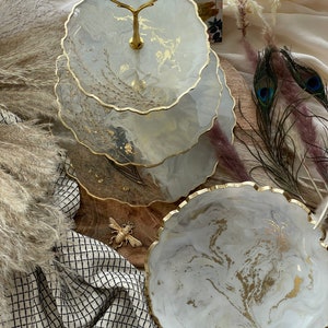 May include: A three-tiered serving tray with a gold handle and trim. The trays have a white and gray marbled design with gold accents. The tray is surrounded by decorative elements, including peacock feathers and dried plants.