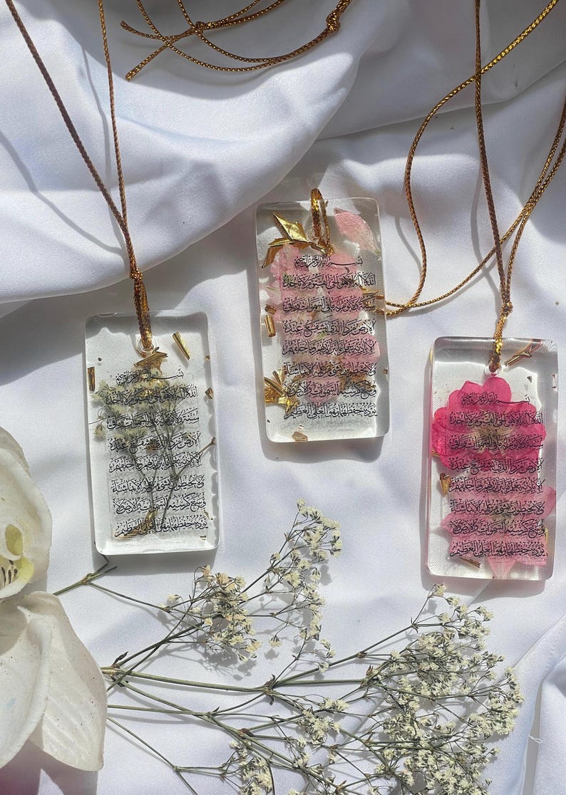 May include: Three rectangular resin pendants with gold chains. Each pendant features dried flowers and Arabic script. One pendant has white flowers, another has pink flowers, and the third has pink flowers. The pendants are displayed on a white fabric.