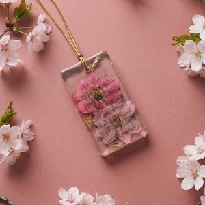 May include: A rectangular pendant with a pink flower encased in clear resin, suspended from a gold chain. The pendant has Arabic script. Pink cherry blossoms surround the pendant on a pink background.