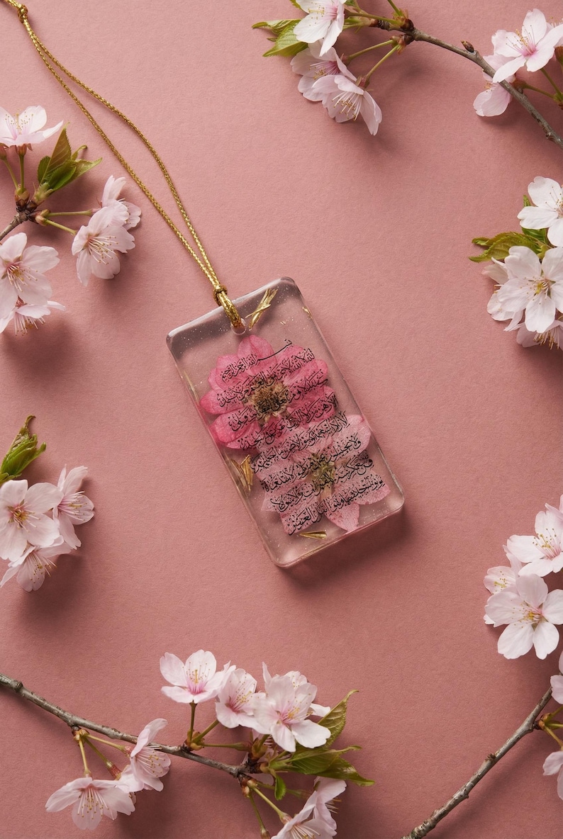 May include: A rectangular pendant with a pink flower encased in clear resin, suspended from a gold chain. The pendant has Arabic script. Pink cherry blossoms surround the pendant on a pink background.