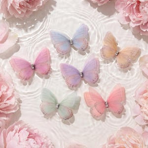 Könnte beinhalten: Sechs transparente Schmetterlingsornamente in Pastelltönen wie Blau, Pfirsich, Rosa, Lila, Mint und Koralle, jeweils mit einem kleinen silberfarbenen Kristallakzent in der Mitte, arrangiert zwischen großen, zartrosa Pfingstrosen im Wasser.