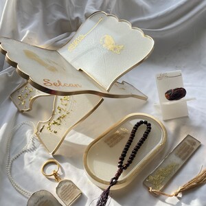 May include: A set of Islamic prayer items including a white prayer stand with gold accents, a white tray with a brown prayer bead necklace, a white bookmark with gold accents, and a white keychain with gold accents. The prayer stand has the word "Selcan" written on it.