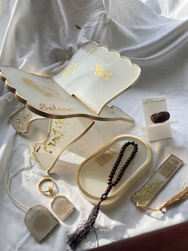 May include: A set of Islamic prayer items including a white prayer stand with gold accents, a white tray with a brown prayer bead necklace, a white bookmark with gold accents, and a white keychain with gold accents. The prayer stand has the word "Selcan" written on it.