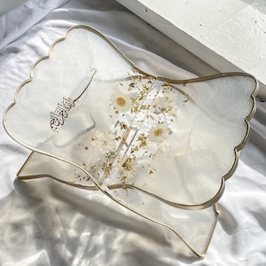 May include: A clear resin book stand with a gold trim and a floral design. The stand is decorated with white flowers and gold leaf. The Arabic word "Allah" is written in gold on the stand.