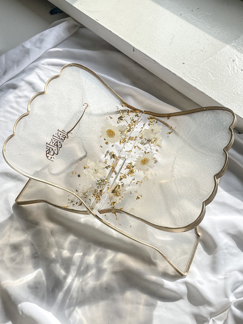 May include: A clear resin book stand with a gold trim and a floral design. The stand is decorated with white flowers and gold leaf. The Arabic word "Allah" is written in gold on the stand.