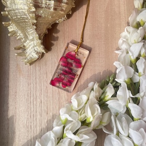 May include: A gold chain necklace with a rectangular pendant. The pendant is clear with a pink floral design. A gold seashell and white flowers are in the background.