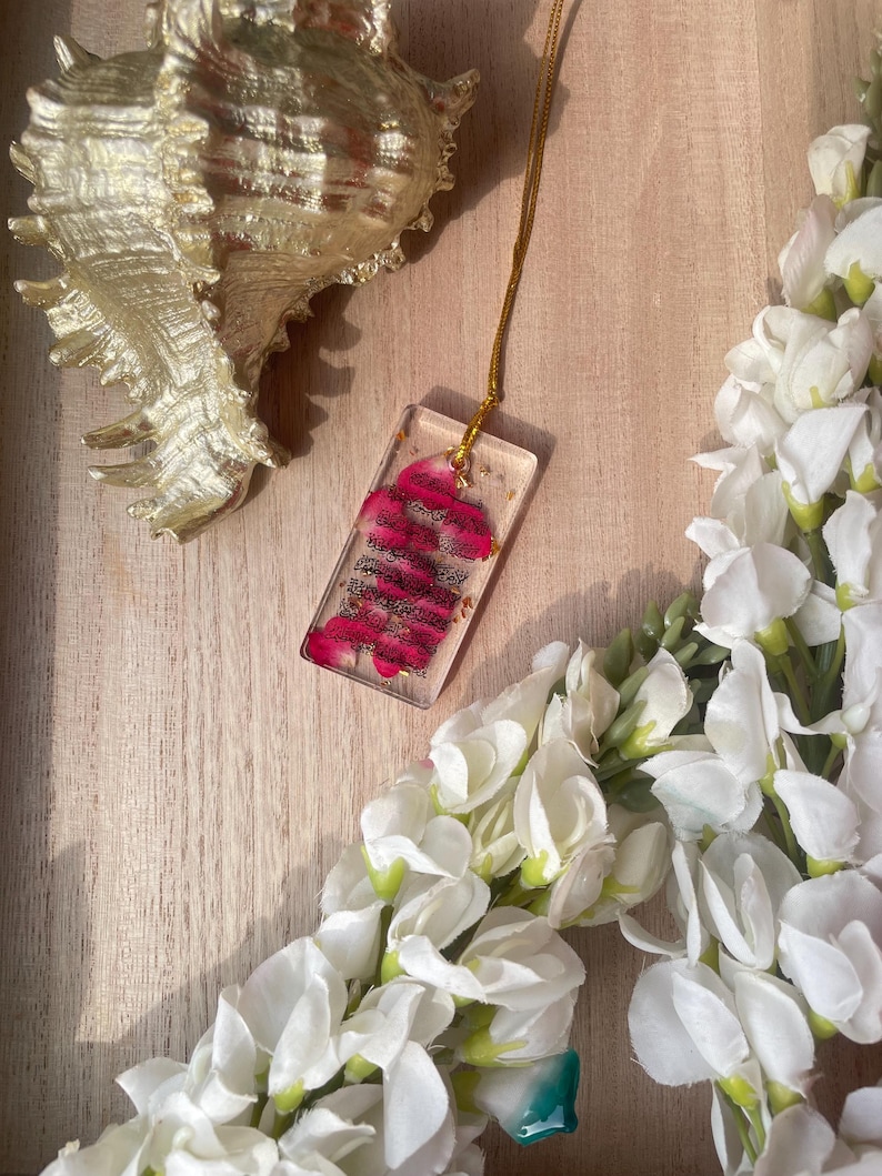 May include: A gold chain necklace with a rectangular pendant. The pendant is clear with a pink floral design. A gold seashell and white flowers are in the background.