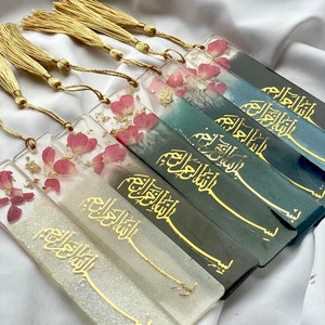 Könnte beinhalten: Ein Set aus sechs Lesezeichen mit goldenen Quasten. Jedes Lesezeichen hat einen anderen Hintergrund in Farbe mit goldener Kalligraphie und getrockneten rosa Blütenblättern. Die Kalligraphie lautet "Bismillah ar-Rahman ar-Rahim", was übersetzt "Im Namen Allahs, des Allerbarmers, des Barmherzigen" bedeutet.
