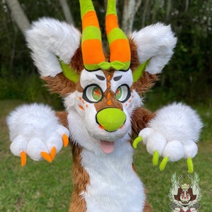 May include: A brown and white furry creature with green and orange horns. The creature has large green eyes and a bright orange tongue sticking out. The creature is wearing a green and orange striped collar. The creature is standing in front of a green grassy background.